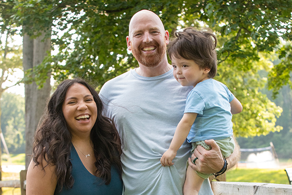 A woman with wavy hair smiles alongside a bald man with a beard holding a toddler with brain cancer.