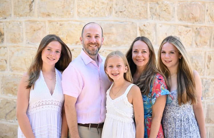 A family of a father in a pink shirt, a mother in a floral dress, and three girls in dresses smile at the camera.
