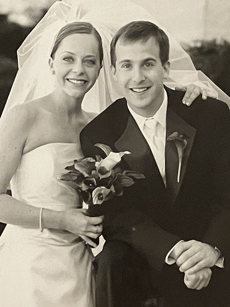 A woman in a strapless white wedding dress and a man in a black tux smile at the camera on their wedding day.