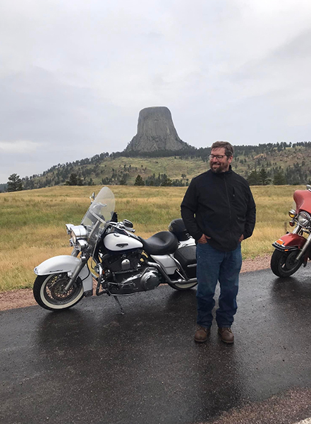 A man in a black jacket walks away from his black-and-white motorcycle with a mountainous landscape in the background.