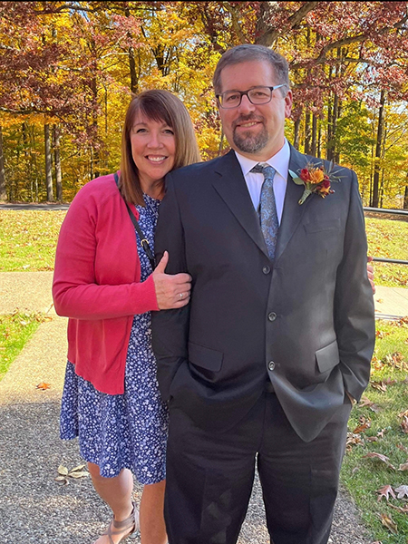 A woman in a pink sweater and dress holds the arm of a man in a suit. The fall foliage is behind them.