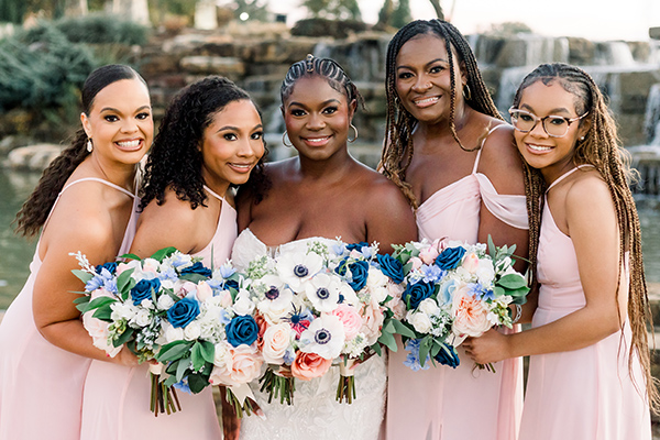 A bride poses with her four bridesmaids in light pink dresses as they hold flowers and look at the camera.