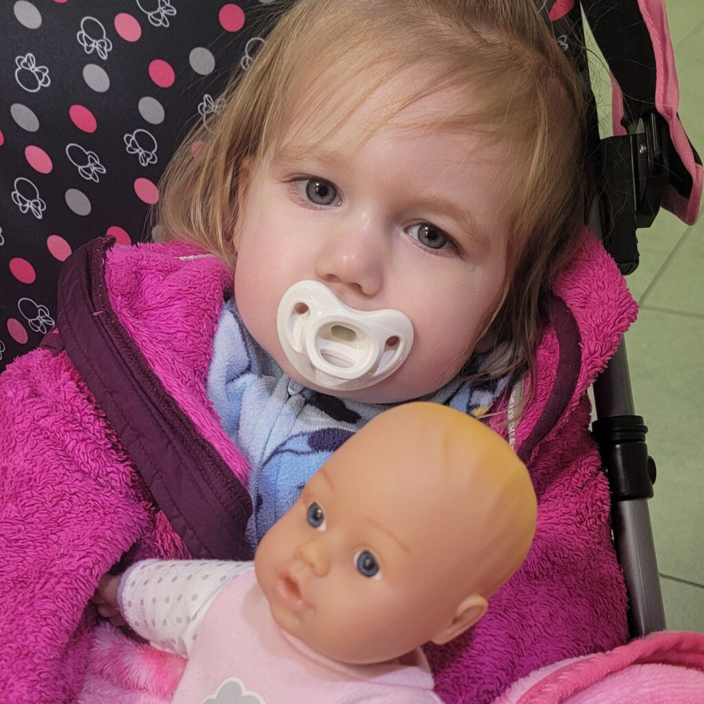 A toddler girl sits in a pink stroller with a pacifier in her mouth and doll on her lap.