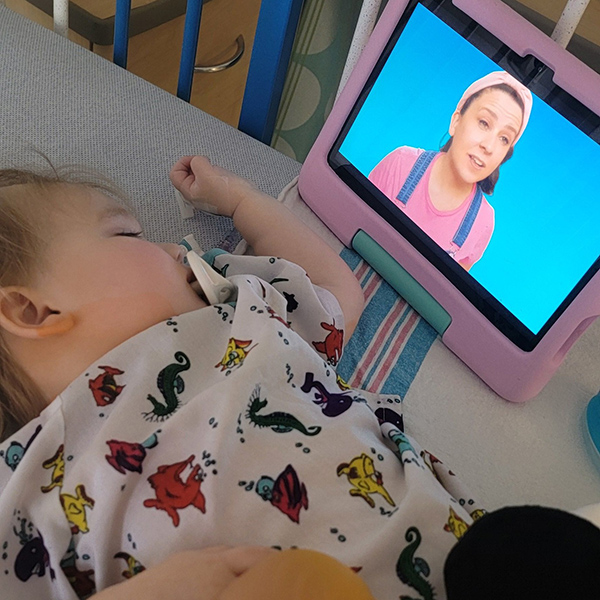 A toddler with grade 1 diffuse astrocytoma with a MYB alteration in a hospital gown and bed watches Ms. Rachel.