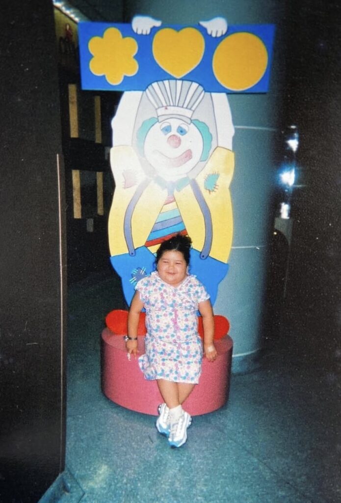 A young child in a hospital gown stands in front of a clown poster.