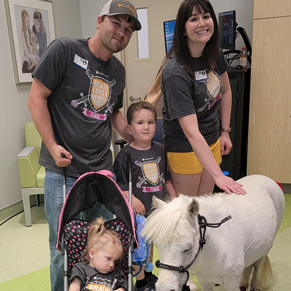A family is in a pediatric hospital. The mom pets a miniature pony, while the dad and young son stand, looking at the camera. A young toddler girl with diffuse astrocytoma with a MYB alteration sits in a stroller.