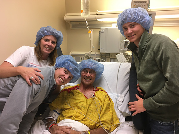 A man with glioblastoma in a yellow hospital gown smiles in a hospital bed with his wife and two teenage boys standing next to his hospital bed before his craniotomy.