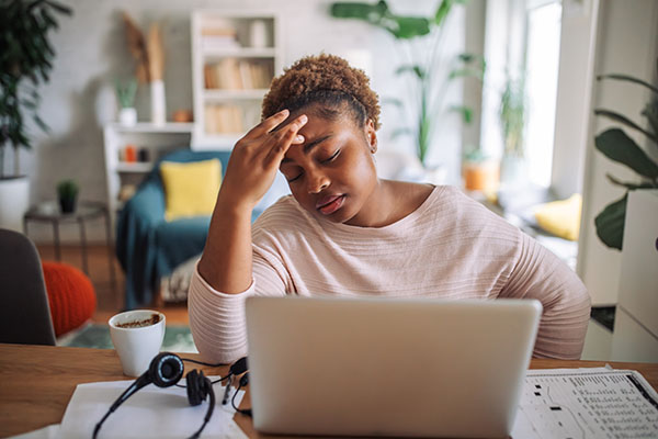 A woman, who is returning to work after a brain tumor diagnosis, is seated at a home office desk in front of a laptop, her hand on her head in a gesture of concern or stress. 