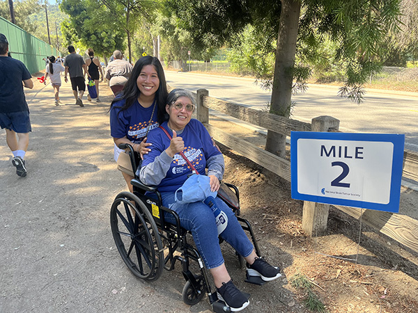 A young adult daughter pushes her mother's wheelchair at a 2-mile marker. They found community through NBTS at the Southern California Brain Tumor Walk.