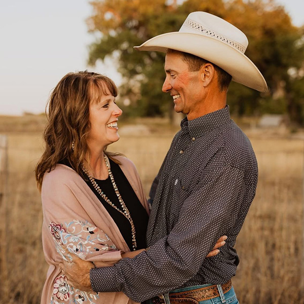 A couple stand in a field. The husband wears a gray button down shirt and cowboy hat, smiling down at his wife who is in a black shirt and pink cover.