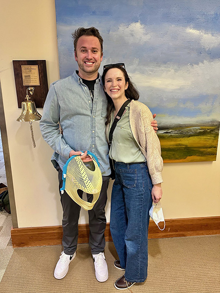 A man holding a radiation mask has his arm around his wife's shoulder. He stands next to the bell, where he rang after his final day of radiation therapy.