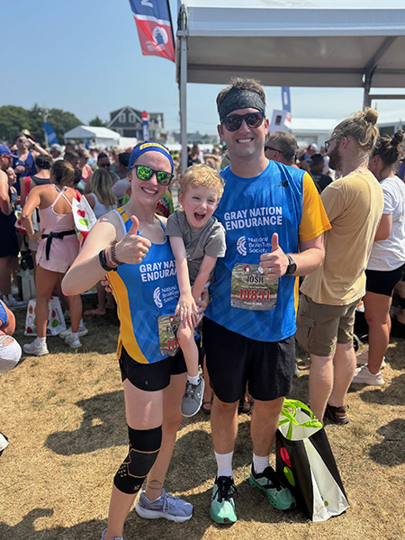A woman and man in Gray Nation Endurance shirts hold their thumbs up after the Falmouth Road Race. Their son is in the woman's arms.