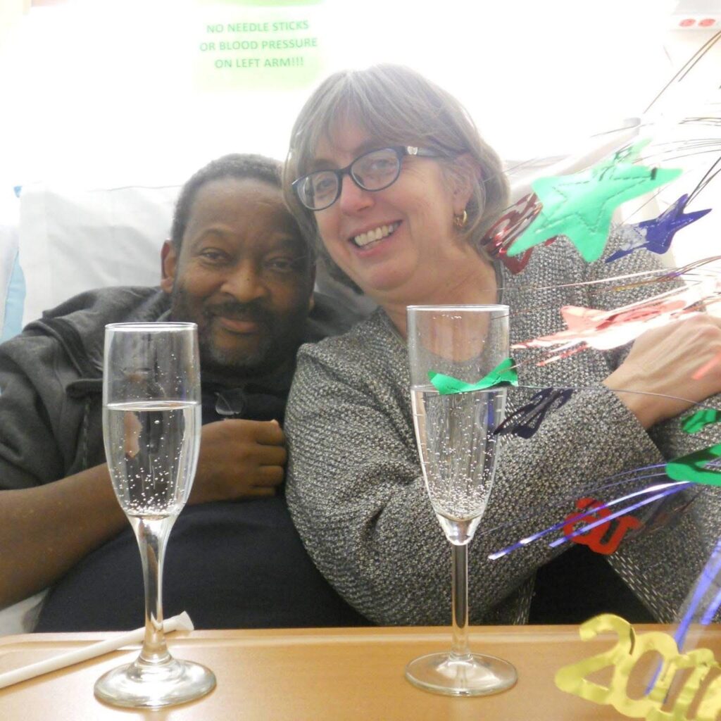 A Black man and a white woman sit in a hospital bed with sparkling juice in front of them as they celebrate New Year's Even.