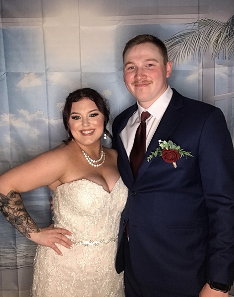 A woman in a wedding dress with a forearm tattoo poses next to her groom in a tuxedo and red tie on the night of their wedding.