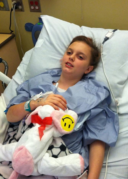 A young girl rests in a hospital bed after being diagnosed with craniopharyngioma brain tumor. She has her hand resting on a white stuffed animal.