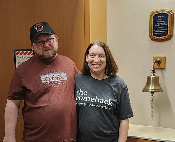A man and woman stand next to a bell that is rung at the end of radiation. The woman's t-shirt reads, "The Comeback is stronger than the setback."
