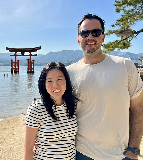 A man in a white t-shirt stands next to a woman in a black and white striped t-shirt on a beach with water, mountains, and an Asian structure in the water behind them.