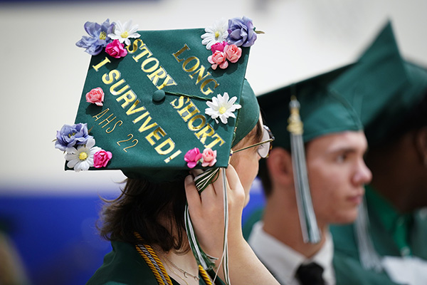 A teenager adjusts the tassle of her graduation cap, which has gold stickers on it that read, "Long story short, I survived!"