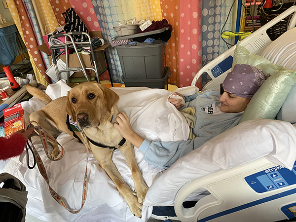A female teenager rests in a hospital bed while petting a therapy dog. She wears a bandana and eye patch.