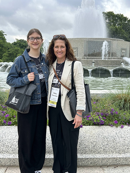 A young adult female in a jean jacket and black pants stands next to her mom, a woman in a cream blazer and black pants, in front of a fountain in D.C. They were in town to advocate for the BRAIN Act.