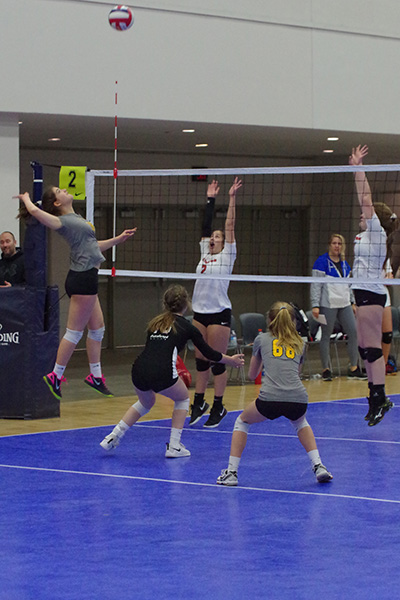 Teenage girls play volleyball on a blue court. One girl jumps to try to spike the ball and two jump to try to block it.