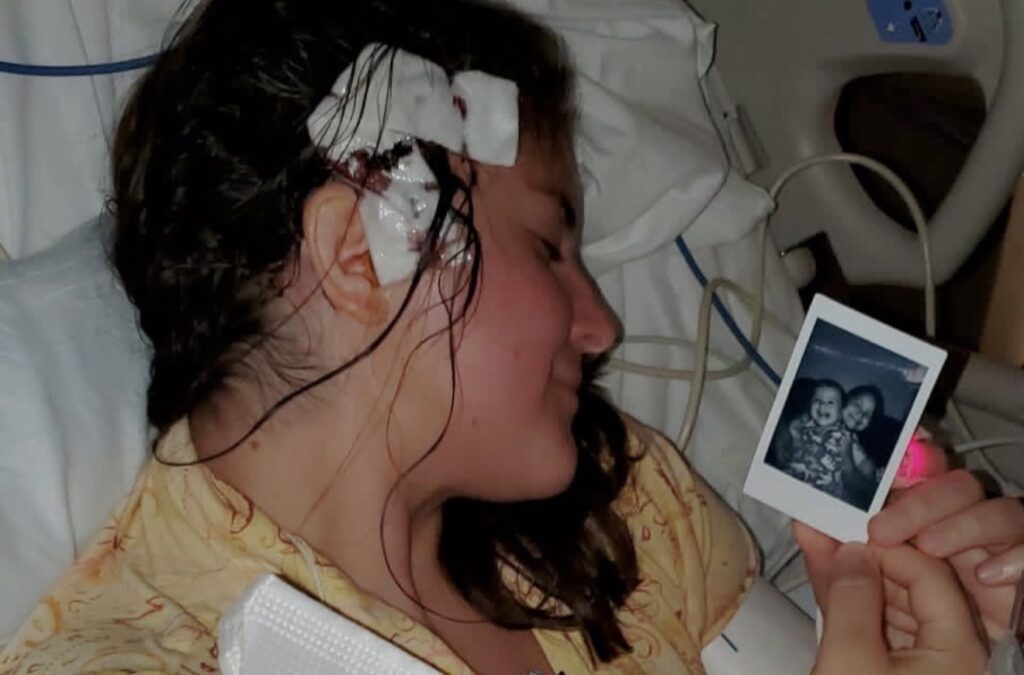 A woman with a bloody bandage on the right side of her head looks at a Polaroid picture of her two little kids while sitting in a hospital bed.