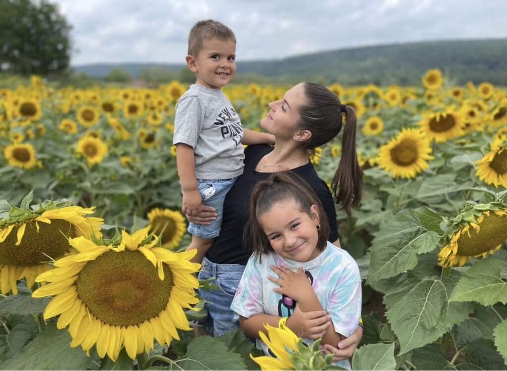 A brunette woman with a ponytail stands in a tall sunflower field, holding her son and her arm wrapped around her daughter standing in front of her.