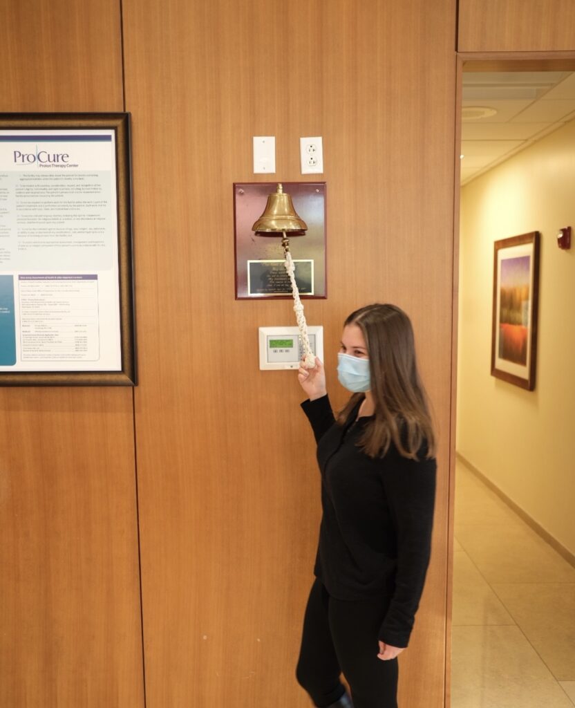 A brunette woman in a mask rings a radiation bell after completing treatment for a brain tumor.