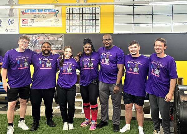 Seven adults clad in purple Lifting for Lavi shirts stand in a gym for a fundraiser benefiting the National Brain Tumor Society.