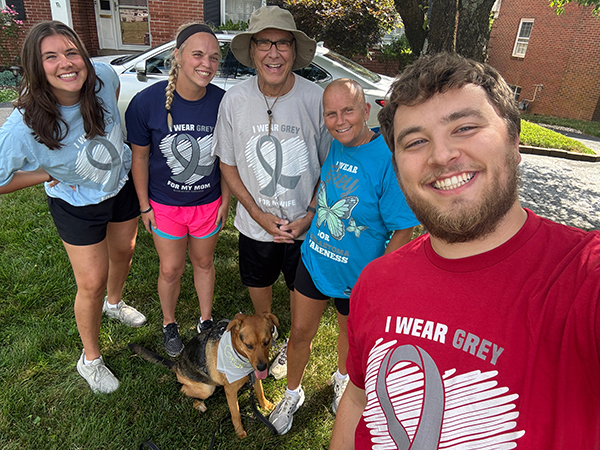 A family of five — three young adults and two older parents — pose for their camera in different "I wear grey for..." t-shirts. The older woman has a bald head from treatment during a glioblastoma clinical trial.