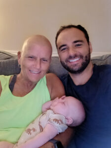 A woman in a sleeveless green top and with a bald head from glioblastoma treatment holds her grandchild while sitting next to a young adult male on a couch.