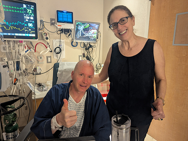 A man sits in a hospital room in a hospital gown and blue robe, giving a thumbs up, while a woman stands next to him in a black sleeveless shirt.