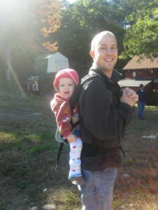 A toddler in a pink hat and jacket sits in a carrier on the back of her father in a black jacket and jeans outside.