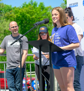 A teenage girl in an NBTS event t-shirt speaks at the National Brain Tumor Ride with her father standing in the background watching her speak.