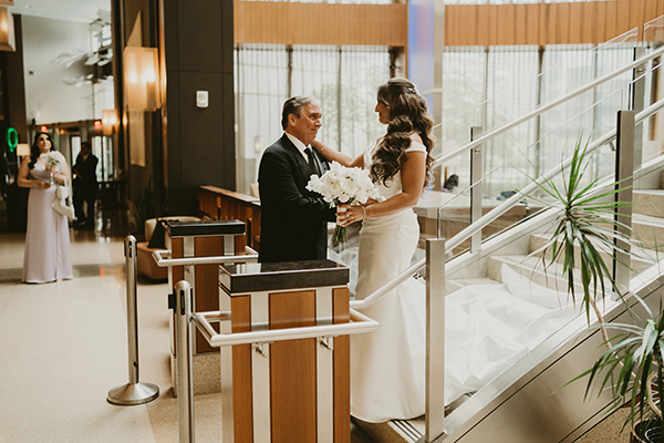 A bride with long brain hair in a wedding dress walks down the stairs as her father, clad in a dark suit, greets her at the bottom of the stairs.