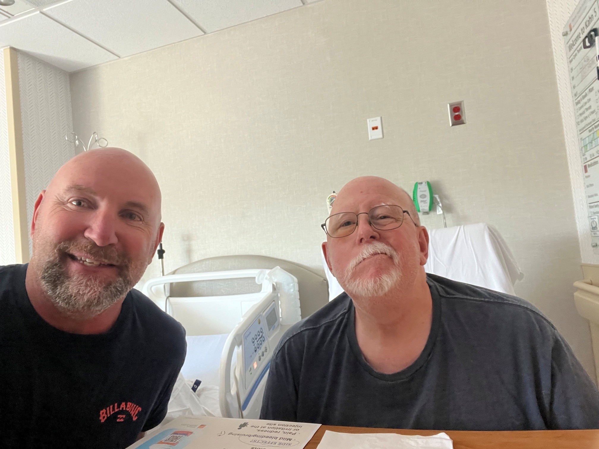 Two men sit in a hospital room. The older man on the right has a crooked smile and glasses. His adult son sits to the left.