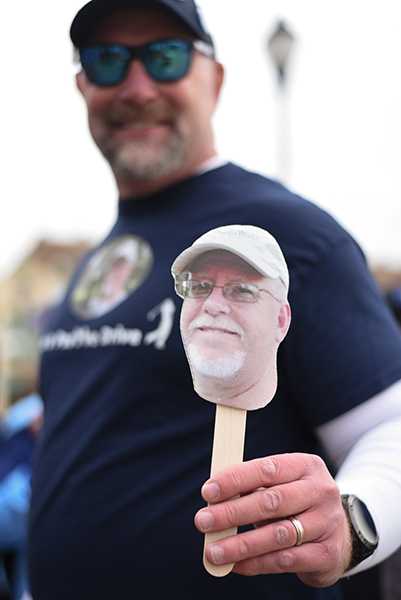 A man, who is blurry in the background with a hat and sunglasses, holds a popsicle stick with a photo of his dad, who had passed from glioblastoma, at the top of the stick.