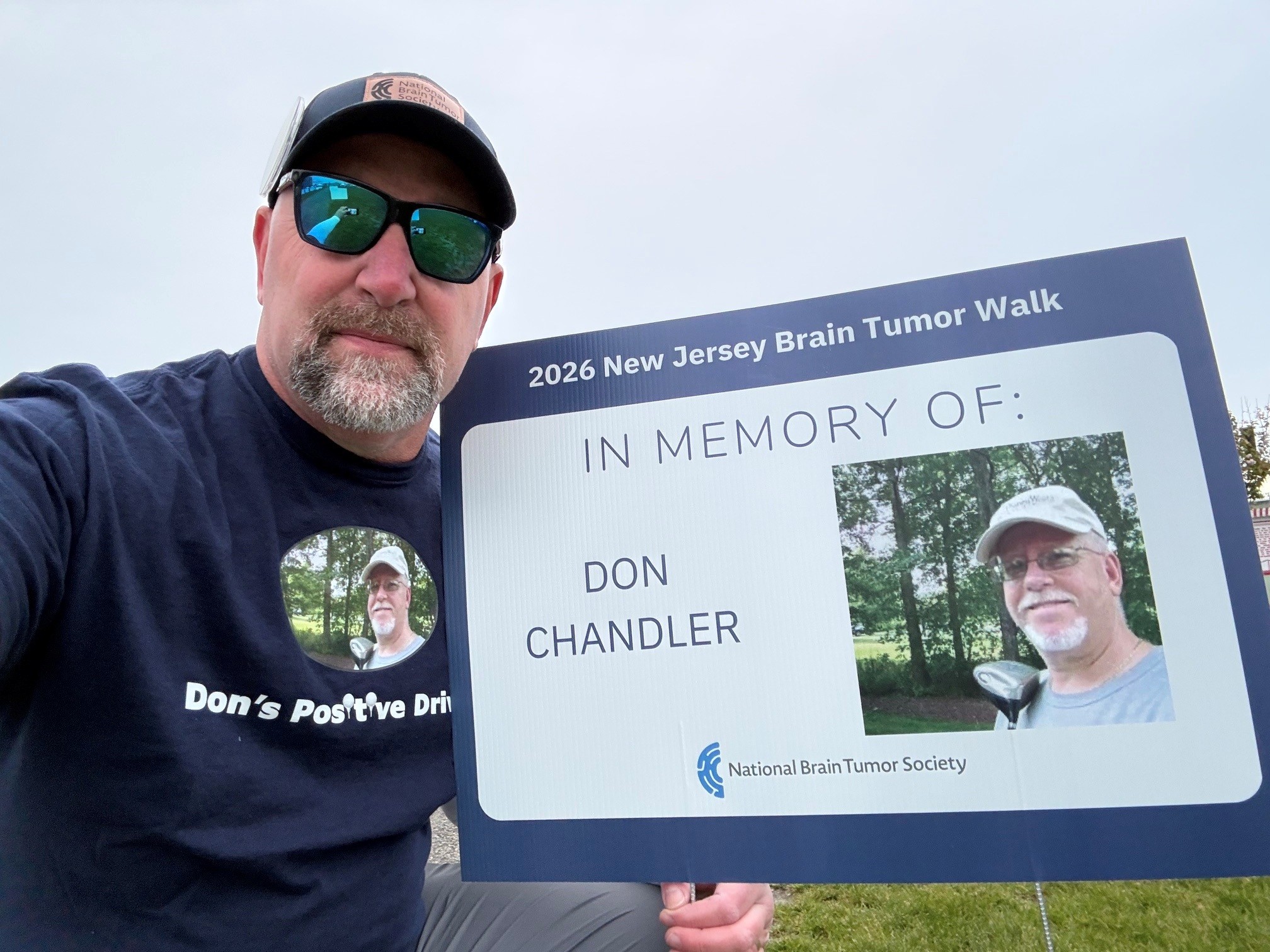 A man in a hat, sunglasses, and a Don's Positive Drive t-shirt holds up an "In Memory of Don Chandler" sign from the New Jersey Brain Tumor Walk.