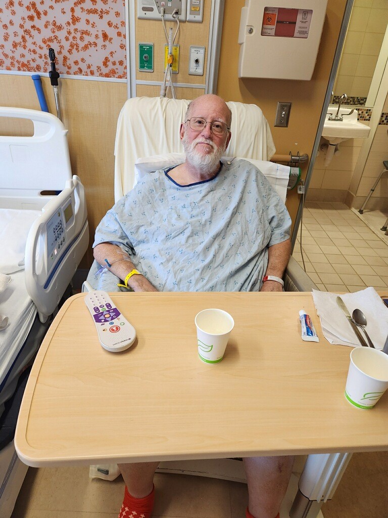 A man with a white beard and wearing a hospital gown sits in a hospital chair with a hospital table in front of him.