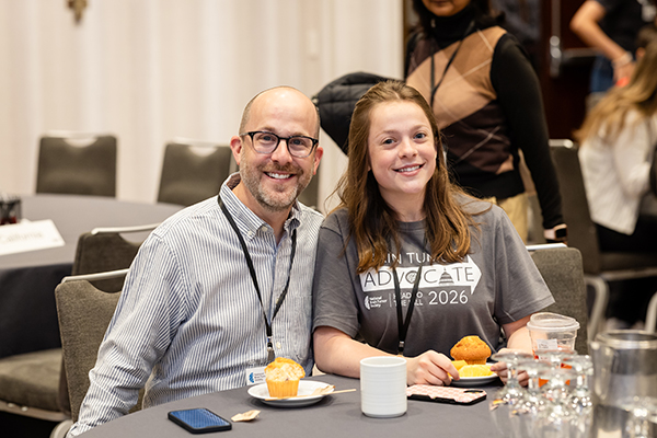 A father in a long-sleeve shirt sits next to his teenage daughter in a brain tumor advocate shirt at a breakfast table.