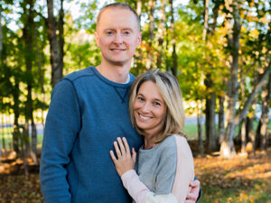 A woman puts her hand on her significant other's chest. The man in a blue sweater stands next to her in a wooded area.