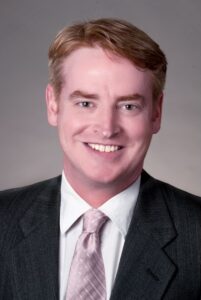A man in black suit with a light pink tie and white shirt looks at camera for headshot photo.