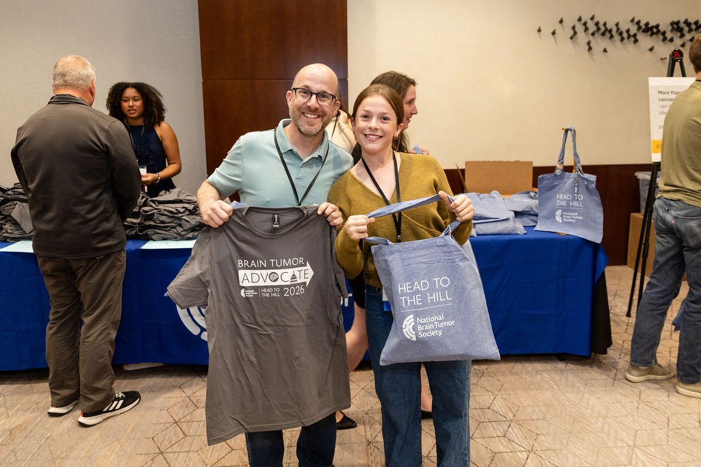 A father holds up a brain tumor advocate t-shirt standing next to his teenage daughter holding up a Head to the Hill bag.