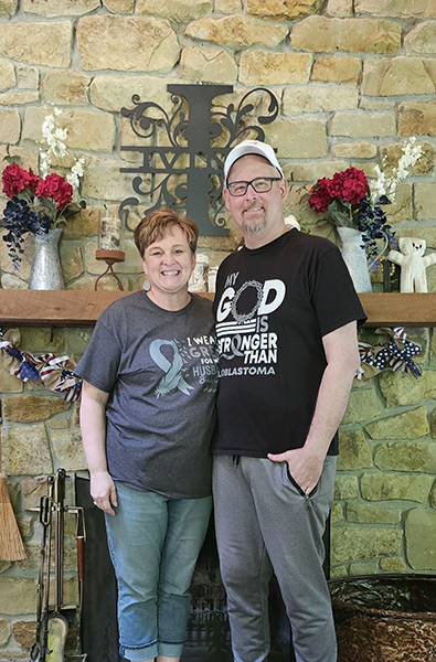 A woman and man stand in front of a mantle with roses behind them. They're each wearing shirts that read, "My God is Bigger Than Glioblastoma."