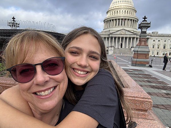 A teenage girl in a dark t-shirt wraps her arms around her mom's neck, both wearing smiles, as they stand in front of the Capitol as pediatric brain tumor advocates.