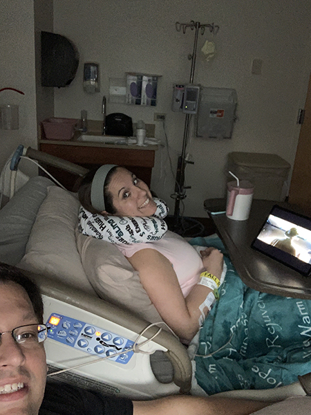 A woman smiles at the camera while sitting in a hospital bed with a neck pillow.