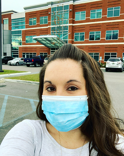 A woman stands in front of a health building wearing a blue surgical mask during the COVID pandemic.
