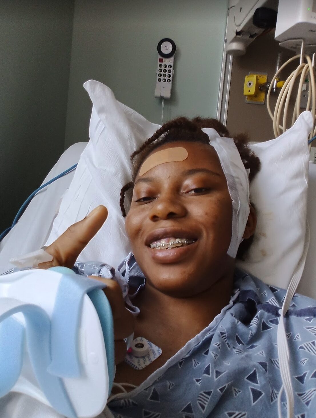 A woman with braces smiles in a hospital bed with a bandaid and a surgical bandage on her face.