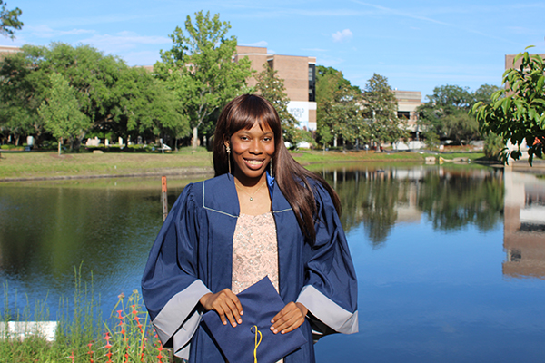 A Black woman in a dress and a blue graduation gown holds her cap in front of a pond.