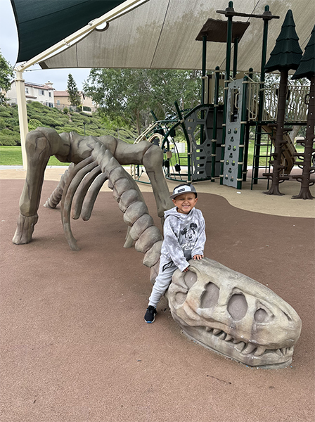 A young boy in a Mickey sweatshirt, sweatpants, and hat sits on top of a playground dino skeleton.